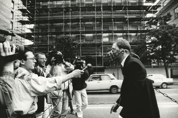 Reporters holding microphones out to a man in a mask.